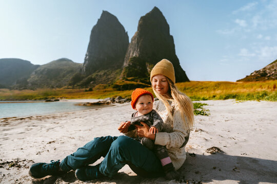 Family lifestyle mother and infant baby travel together sitting on the beach in Norway, outdoor vacation bonding mom with child having fun, candid emotions parent with kid explore Helgeland islands