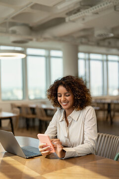 Vertical portrait of american business woman holding mobile phone near laptop in coworking office workspace, using app. In 30s, female founder works as analyst, advisor, agent, banker, broker