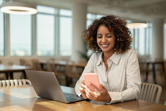 Young multiracial business woman holding mobile phone near laptop in coworking office workspace, using app. In 30s, female manager works as executive, entrepreneur, owner, ceo, boss. Copy space