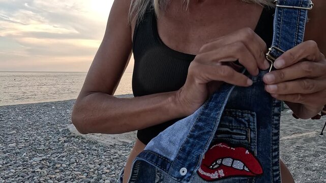 Woman fastening denim overalls strap on pebble beach at sunset. Medium closeup shot.