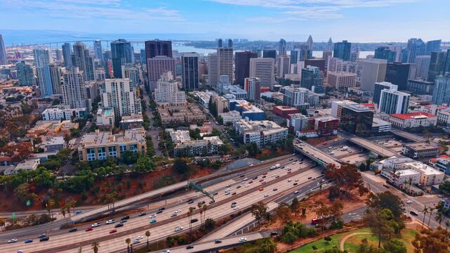 San Diego skyline beyond freeway traffic and parkland. Cars cross parallel lanes below rows of towers in San Diego, with trees and grass in the foreground.