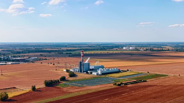 Factory chimney and solar panels beside harvested farmland. A white plume drifts above an industrial building between crop fields and a rectangular module array.