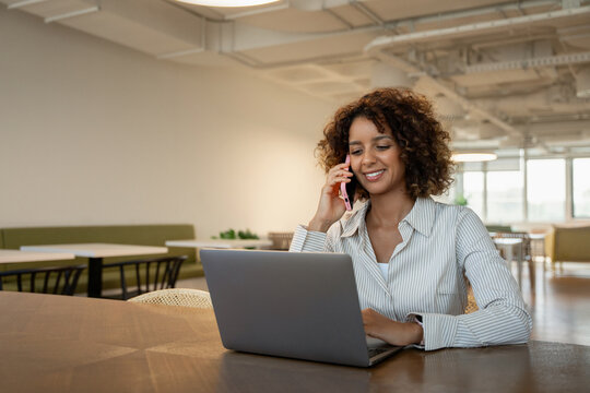 Smiling latin business woman talking on mobile phone in coworking office workspace, having client call. In 30s, female owner works as ceo, boss, leader, specialist, consultant, director. Copy space