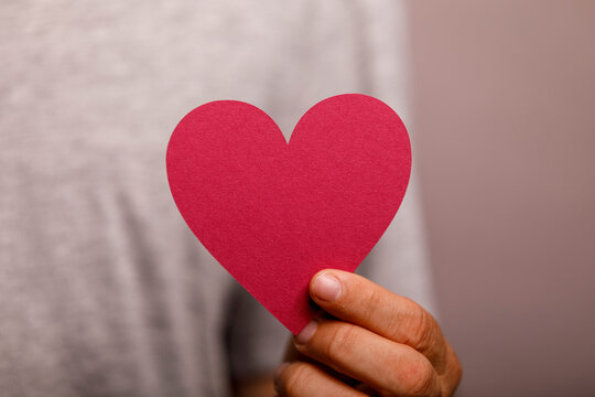 A person's hand offers a red paper heart, conveying love, charity, or support.