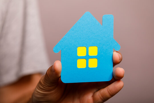 A person's hand holds a small blue paper house, symbolizing homeownership and real estate.