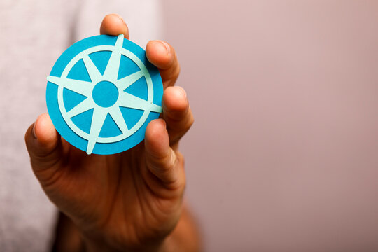 A person's hand holds a blue paper cutout resembling a dharma wheel or ship's wheel.