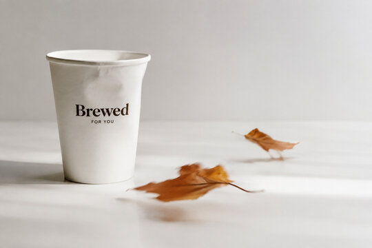 Disposable paper cup with deformed rim and dried autumn leaves in soft breeze, bright studio