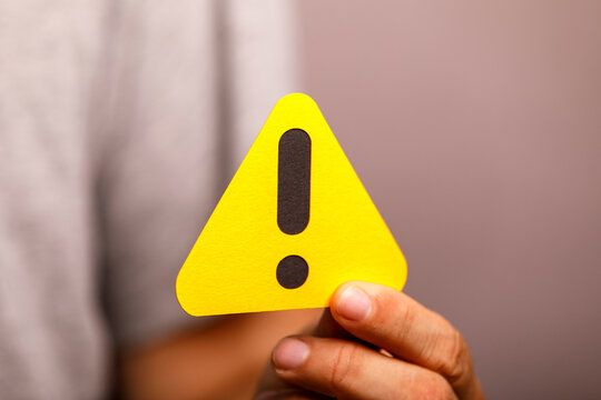 A hand holds a bright yellow triangular warning sign with a black exclamation mark.