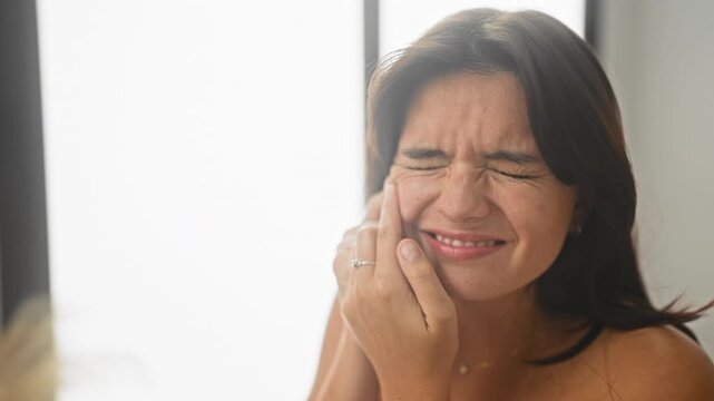 Woman pressing cheek with hand and wincing, bare shoulder visible, candid portrait in studio near bright window showing dental pain; toothache pain.