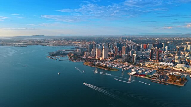 Boats leaving wakes below the San Diego waterfront skyline. Small vessels cross San Diego Bay beneath towers and marina docks, tracing white lines across the water.