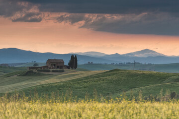 Obraz premium Traditional stone farmhouse standing on rolling green hills in Tuscany at sunset, framed by distant mountains and a dramatic evening sky. Ideal for travel and agritourism concepts.
