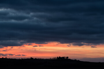 Fototapeta premium Silhouette of cypress trees on a hill ridge under dark moody storm clouds and vibrant orange sunset glow. Ideal background for travel and serenity concepts.