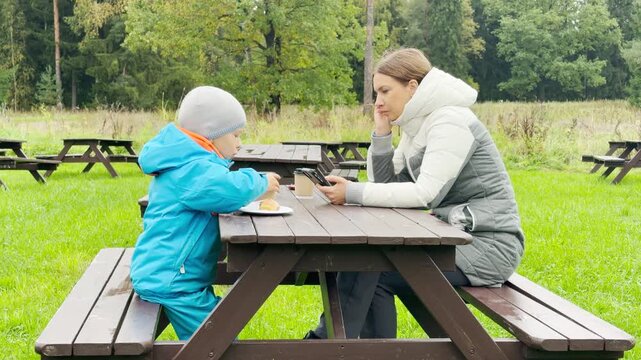 A mother and child sit together enjoying a peaceful snack at a wooden table. They rest comfortably surrounded by green nature in Pavlovsk Park, Saint Petersburg.