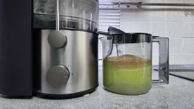 Electric juicer extracting fresh green juice filling transparent pitcher in kitchen, time-lapse
