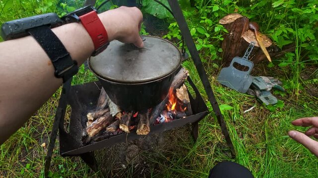Man opening cauldron lid with boiling soup over campfire for outdoor camping lifestyle