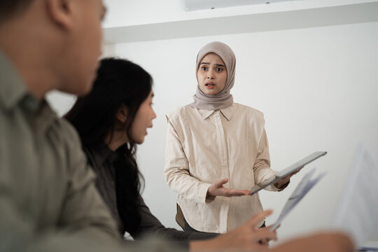 Young woman in hijab gestures with tablet while speaking to coworkers in bright minimalist office, with blurred colleagues in foreground for engaging teamwork discussion.