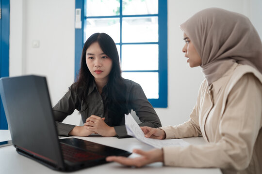 Two professional women review documents during focused office discussion beside open laptop in bright modern workspace, with one woman wearing hijab and other listening attentively across desk
