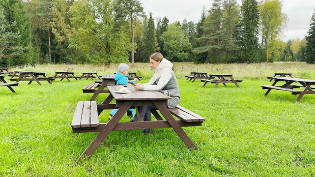 A mother and child sit together at a picnic table. They enjoy a peaceful break on a lush green lawn in Pavlovsk Park, Saint Petersburg. This quiet moment brings warm happiness.