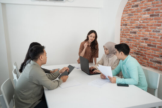 Diverse coworkers gather around laptop and printed documents during focused project discussion in bright office with white table, smartphone, and brick accent wall, showing teamwork and planning.