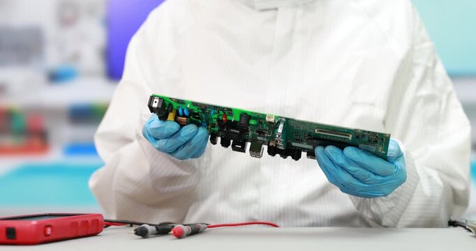 Technician in clean protective clothing and blue gloves examines a green circuit board with testing tools on a laboratory workbench.