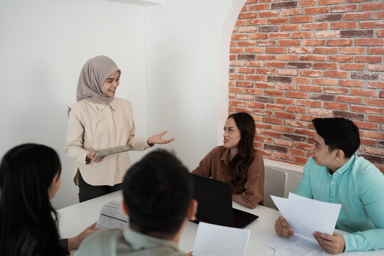 Smiling presenter in hijab shares ideas with diverse coworkers during workplace discussion, standing by laptop and documents near brick wall in modern office for team communication and planning.