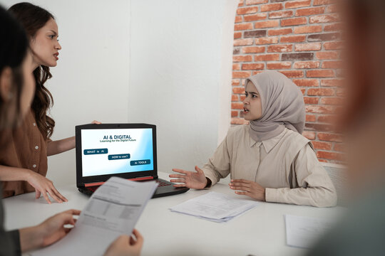 Two women in office discussion over laptop presentation about digital learning, with one presenter speaking and colleague in hijab reacting beside papers on table against brick wall backdrop