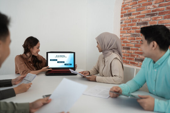Diverse coworkers review printed materials and discuss digital training around laptop in bright meeting room, with brick wall backdrop, relaxed smiles, and collaborative office atmosphere.