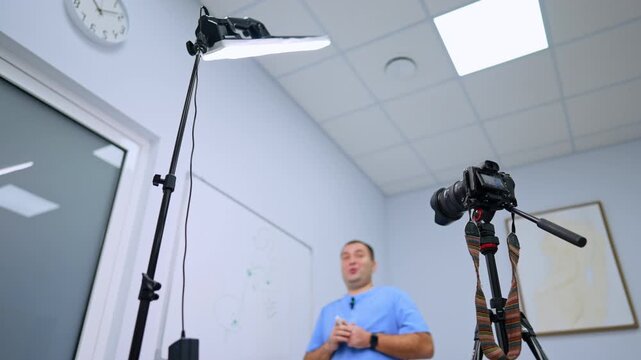 Low angle view of a filming setup and medical presenter. A camera and panel light rise above a man in blue scrubs beside a whiteboard and wall clock.