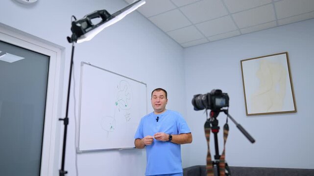 Medical presenter explaining a urinary system diagram on camera. A man in blue scrubs holds a marker beside a kidney sketch, with a tripod camera and panel light in front.
