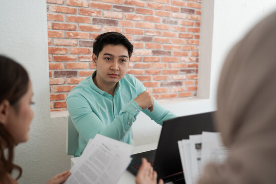 Young man in mint shirt sits at desk during team discussion, listening to colleagues as papers and laptop sit nearby in bright modern office with brick wall background.