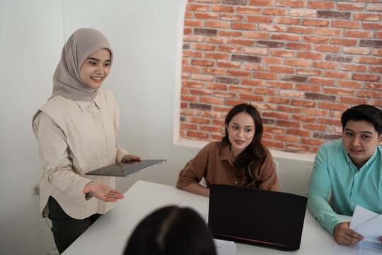 Woman in hijab stands beside desk holding tablet as colleagues sit with laptop and documents, collaborating during discussion in bright office with brick wall backdrop.