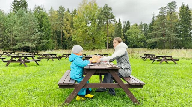 A mother and child sit together at a wooden picnic table in Pavlovsk Park, Saint Petersburg. They share a warm meal near the forest, creating a sweet feeling of peaceful joy and love.
