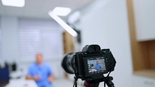 Rear camera monitor showing a medical presenter at a desk. A man in blue scrubs appears on the display while office cabinets and ceiling lights remain blurred.