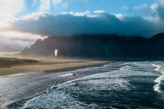 Aerial view of a kitesurfer gliding across the water near the dramatic Vestrahorn mountain, with dark clouds hanging over the beach in Sveitarfelagid Hornafjordur, Iceland.