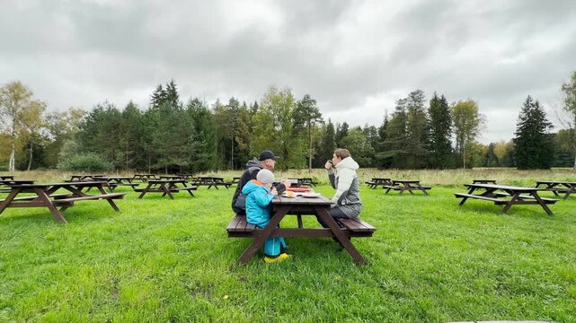 A family rests comfortably at a wooden table, enjoying hot drinks on a green lawn. This cozy gathering brings a warm sense of peace to Pavlovsk Park, Saint Petersburg.