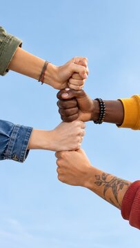 Diverse group of multiethnic friends stacking fists together against blue sky symbolizing teamwork unity and support