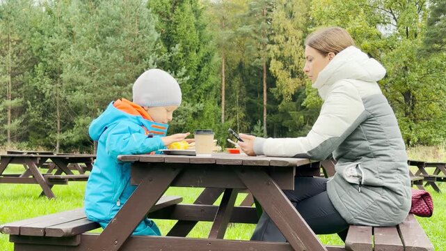 A mother and her child sit at a wooden picnic table to enjoy snacks in Pavlovsk Park, Saint Petersburg. This cozy family resting moment brings a warm feeling of peace and quiet joy.