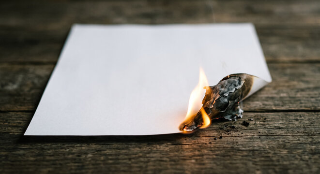 White sheet of paper burning on rustic wooden table with flame curling burnt edge. Concept of small flame, fire, blank sheet, ashes. Copy space.