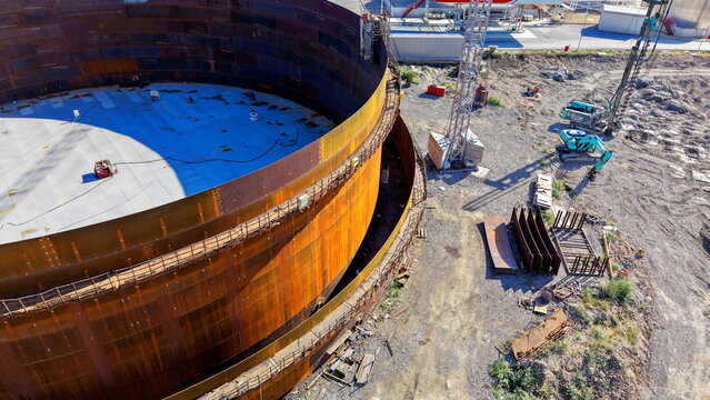 Aerial view of the assembly of a large circular metal oil storage tank at a refinery construction site with heavy machinery and excavators on the ground in Kovin, Vojvodina, Serbia.