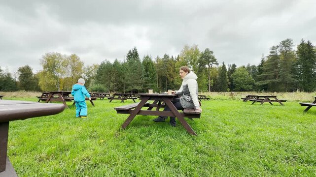 A child walks across a green lawn while a mother rests at a picnic table in Pavlovsk Park, Saint Petersburg. This outdoor scene brings a peaceful feeling of warmth, joy, and serene calm.