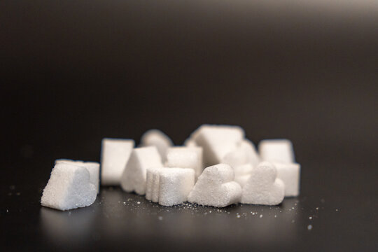 Low angle row of mixed sugar cubes and heart shaped sugar pieces on a dark reflective table with scattered crystals and a soft brown background, concept of sweetness, sugar intake and dessert ingredi