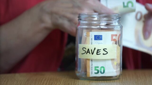 Close-up of a person placing euro cash into a transparent glass savings jar labeled "Saves" on a wooden table.