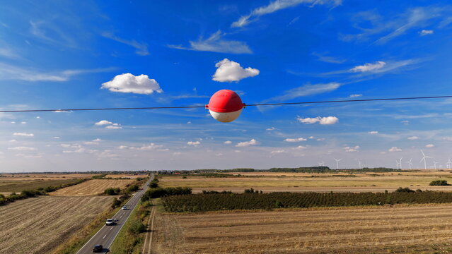 Aerial view of a red and white aerial marker ball on a power line, overlooking a rural landscape with a road, cars, and wind turbines in the distance in Bavaniste, Vojvodina, Serbia.