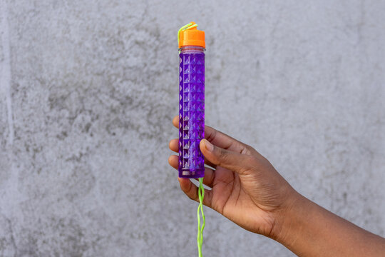 Hand displaying purple plastic bubble maker tube with orange cap outdoors. Fun soap bubble blowing toy stick for kids recreation.