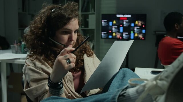 Caucasian young woman wearing headset reading paper document while working in scam call center. Suitable for cybersecurity training and scam awareness
