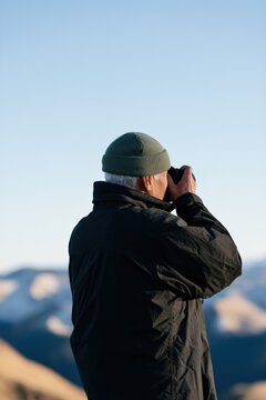 Senior man looking through binoculars in mountain landscape background for travel and outdoor adventure banner and wallpaper