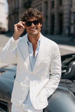 Portrait of a stylish man in a white blazer adjusting sunglasses by a convertible car for lifestyle banner and fashion background