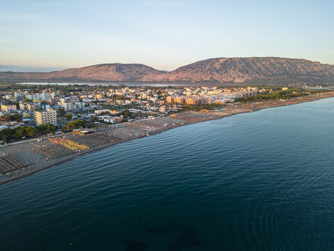 Aerial view of Velipoja Beach with rows of colorful umbrellas on the sandy shore, coastal town buildings, and mountains in the background in Velipoje, Shkoder County, Albania.