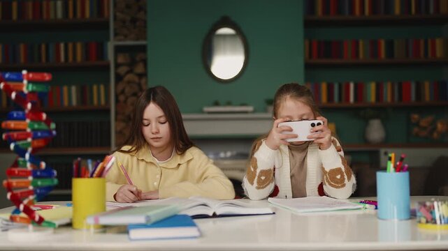 Focused girl does homework while her younger sister procrastinates on phone