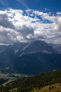 Aerial view of the Dolomites mountain range with dramatic clouds, rugged rocky peaks, and lush green forests in the valley near Rocca Pietore, Veneto, Italy.